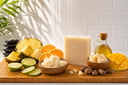 Wooden cutting board with pineapple, oranges, limes, a white soap bar, a bottle of oil, and other items against a white tiled wall.