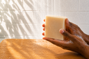 Hand holding a bar of soap on a wooden surface with a tiled wall background