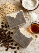 Two bars of soap with coffee beans, oats, and coconut on a wooden surface.