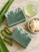 Two green soap bars with aloe vera leaves and aloe vera gel on a wooden surface
