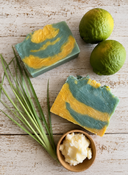 Two bars of green and yellow soap with two limes and a bowl of shea butter on a wooden surface.
