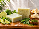 Wooden cutting board with limes, mint leaves, two green soap bars, small wooden bowls of shea butter, mango butter, spinach powder, and other items against a white tiled wall.