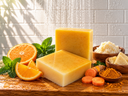 Two rectangular bars of pale yellow soap standing on a wooden surface, surrounded by orange slices, mint leaves, turmeric root, turmeric powder, and bowls of ingredients. Water droplets are falling in the background against a white tiled wall.