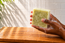 Hand holding a bar of green colored soap on a wooden surface with a tiled wall background