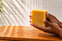 A hand holds a rectangular bar of pale yellow soap under running water, with soap lather visible on the fingers, set against a tiled wall and wooden surface.